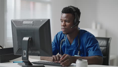 Afro-american-male-doctor-or-nurse-with-headset-and-computer-working-at-hospital-.young-professional-therapist-doctor-consulting-customer-client-using-remote-communication-speaking-on-webcam